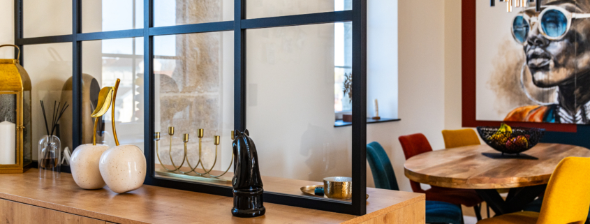 ue d'un espace salle à manger à travers une cloison vitrée à cadre noir posée sur un meuble en bois clair. Au premier plan, deux sculptures de cerises blanches et des objets décoratifs. En arrière-plan, une table ronde avec des chaises colorées (jaune, bleu, rouge terracotta). Une grande toile artistique représentant un visage orne le mur du fond, peint en bleu et terracotta. Une section de mur en pierre ancienne est visible sur la gauche, près d'une fenêtre. Une suspension graphique moderne est accrochée au-dessus de la table.